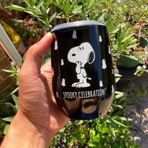 May include: Black stainless steel tumbler featuring Snoopy in a white graphic, surrounded by white ghost illustrations. The tumbler has the text "Spooky Celebration" in white. A fun, festive item for Halloween.