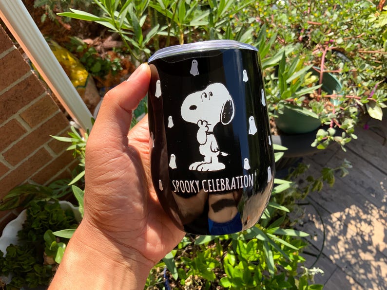 May include: Black stainless steel tumbler featuring Snoopy in a white graphic, surrounded by white ghost illustrations. The tumbler has the text "Spooky Celebration" in white. A fun, festive item for Halloween.