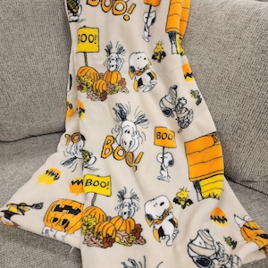 May include: A cream-coloured Halloween throw blanket featuring Snoopy in various costumes, pumpkins, and the word "BOO!" in orange and yellow. The blanket is folded over a grey sofa.
