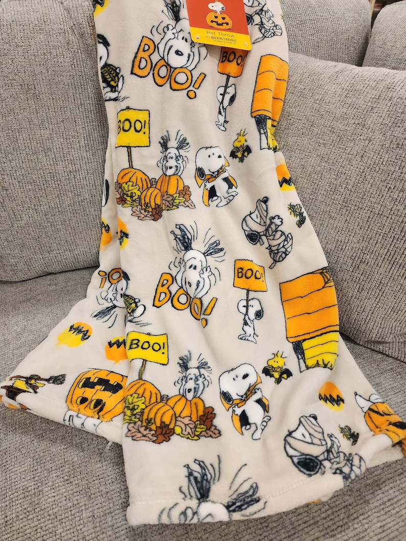 May include: A cream-coloured Halloween throw blanket featuring Snoopy in various costumes, pumpkins, and the word "BOO!" in orange and yellow. The blanket is folded over a grey sofa.
