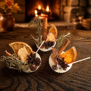 May include: Three white wax melts decorated with dried orange slices, cinnamon sticks, star anise, small pinecones, and sprigs of green rosemary. Each round wax melt features a white wick and is arranged on a dark brown wooden surface.