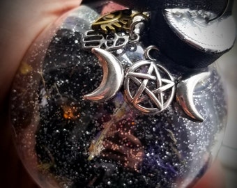 May include: A clear glass ornament filled with black glitter and dried flowers. The ornament is adorned with silver charms including a crescent moon, a pentagram, and a triple moon symbol. A black ribbon is attached to the top, with a gold sun and hand charm.