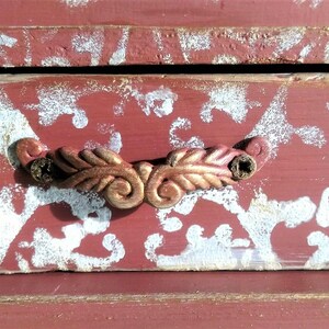 May include: A red wooden box with a white floral pattern and a gold metal handle. The box has a distressed look and is perfect for storing small items.