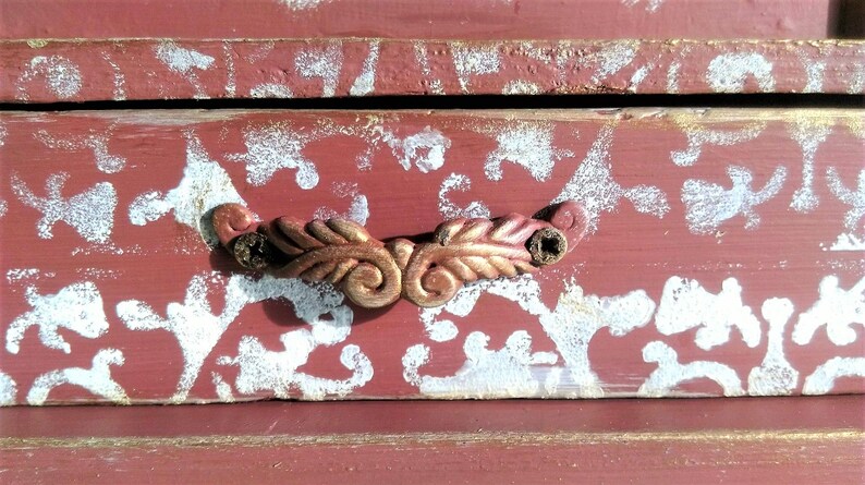 May include: A red wooden box with a white floral pattern and a gold metal handle. The box has a distressed look and is perfect for storing small items.