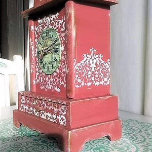 May include: A red wooden clock with a white face and black hands. The clock has a decorative design on the front and sides. The clock is sitting on a white surface with a green design.