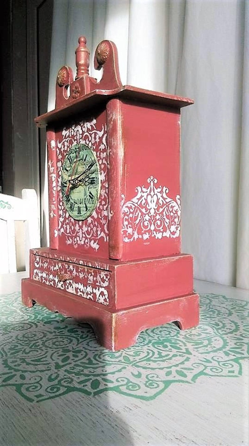 May include: A red wooden clock with a white face and black hands. The clock has a decorative design on the front and sides. The clock is sitting on a white surface with a green design.