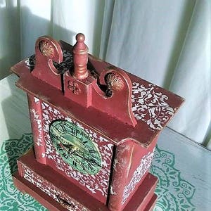 May include: A red wooden clock with a white face and a drawer. The clock has a decorative design on the top and sides. The clock is sitting on a white surface with a green and white pattern.