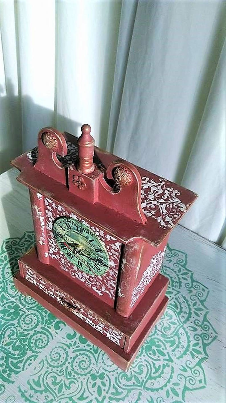 May include: A red wooden clock with a white face and a drawer. The clock has a decorative design on the top and sides. The clock is sitting on a white surface with a green and white pattern.