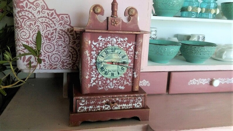 May include: A small, red wooden clock with a white face and black hands. The clock has a decorative drawer and is decorated with a white stencil design.