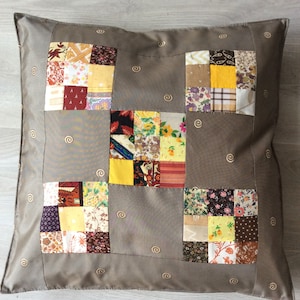 Peut inclure: Coussin carré décoratif avec un motif patchwork. Le centre présente un patchwork de carrés de tissu colorés aux motifs et couleurs variés, entouré d'un tissu gris uni. Le coussin a une bordure grise avec des détails en spirale brodés.