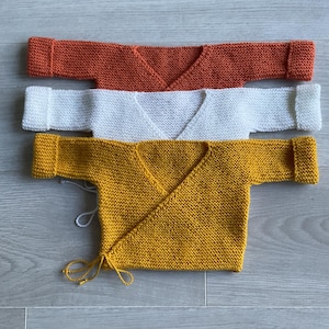 Puede incluir: Tres cárdigans envolventes de punto en naranja, blanco y amarillo. Los cárdigans tienen mangas largas y cuello en V. El cárdigan amarillo tiene cierres de lazo. Se muestran sobre una superficie de madera gris claro.