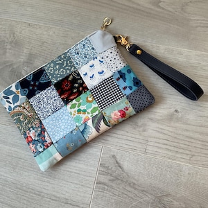 Peut inclure: Une pochette rectangulaire zippée présentant un motif en patchwork composé de divers carrés de tissu bleus, blancs et à motifs floraux. La pochette comprend une dragonne noire amovible fixée à un fermoir et une fermeture éclair dorés.