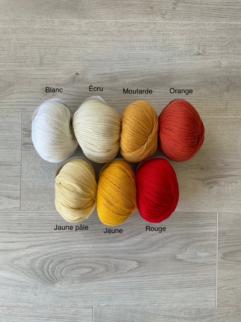 May include: Balls of yarn in various shades of yellow, orange, and red. The yarn is labeled with French words for each color: Blanc, &Eacute;cru, Moutarde, Orange, Jaune p&acirc;le, Jaune, and Rouge.