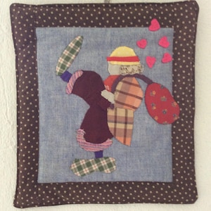 Puede incluir: Una pared de tela marrón con un fondo azul y una figura de patchwork con un sombrero rojo y una bolsa roja y blanca. La figura está rodeada de corazones rosas.