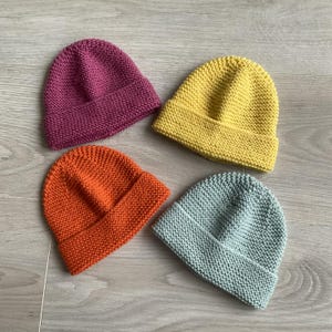 Könnte beinhalten: Vier gestrickte Mützen in verschiedenen Farben: Lila, Gelb, Orange und Hellblau. Jede Mütze hat einen umgeschlagenen Saum und ein strukturiertes Strickmuster. Die Hüte sind auf einer hellen Oberfläche angeordnet.