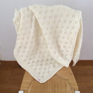 May include: A white crocheted baby blanket with a delicate openwork pattern. The blanket is draped over a chair with a woven seat.