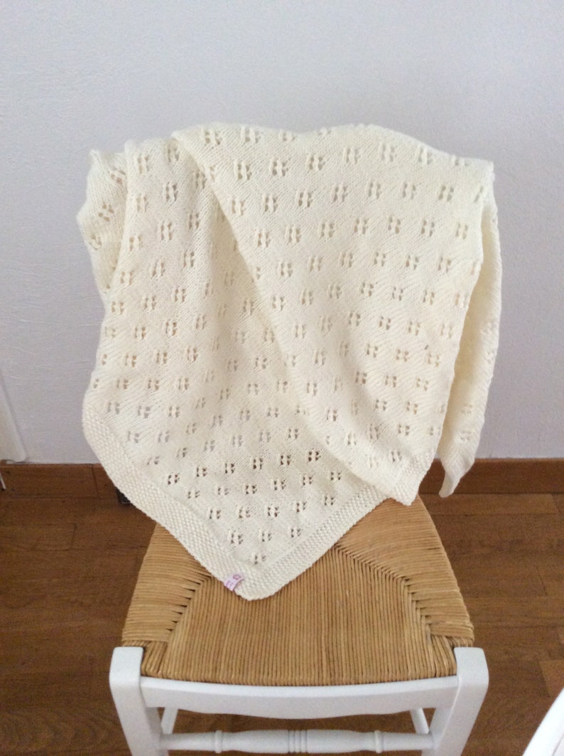 May include: A white crocheted baby blanket with a delicate openwork pattern. The blanket is draped over a chair with a woven seat.