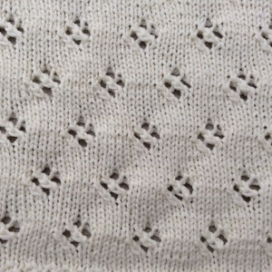 May include: Close-up of a white knitted fabric with a diamond-shaped pattern. The fabric has a delicate, openwork design.