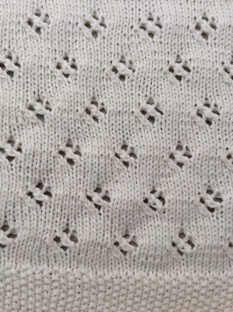 May include: Close-up of a white knitted fabric with a diamond-shaped pattern. The fabric has a delicate, openwork design.
