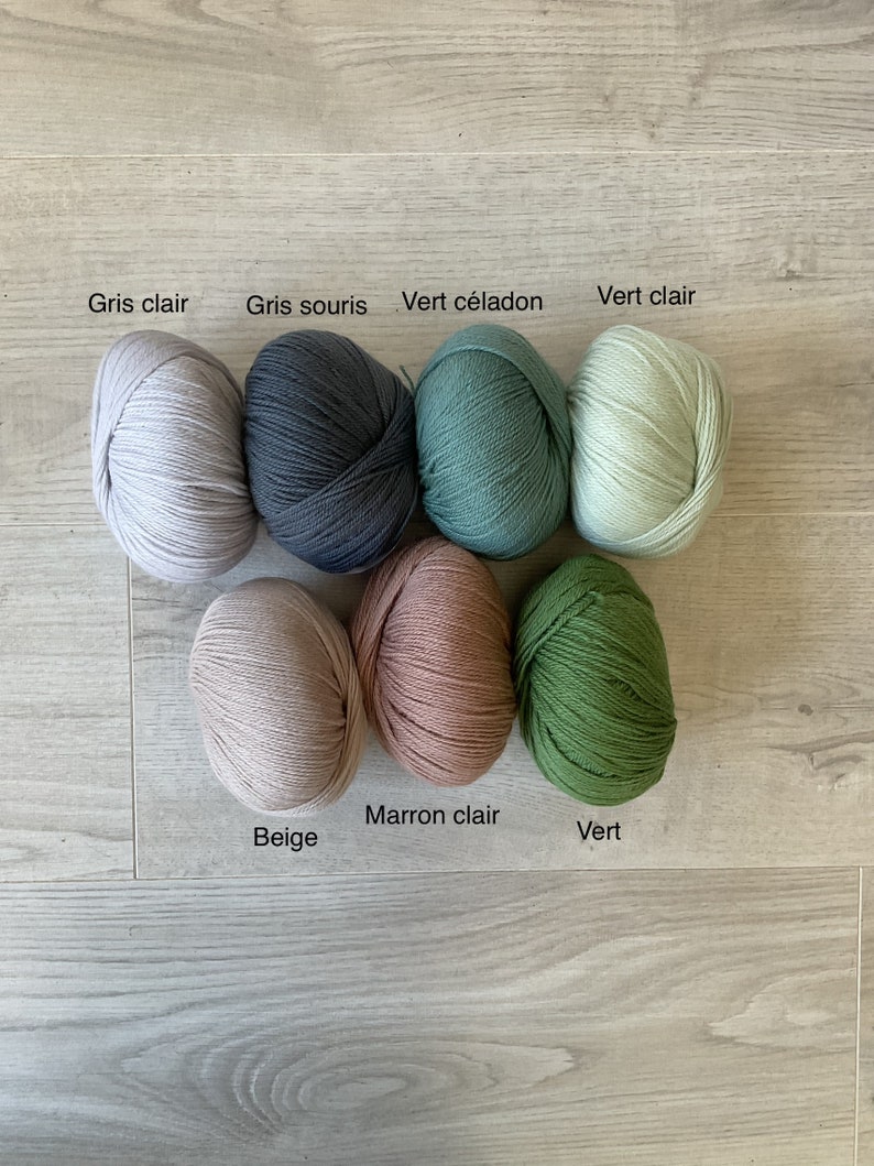 May include: Seven balls of yarn in various colors, including beige, light gray, dark gray, light green, dark green, light brown, and white. The yarn balls are arranged in a circle on a wooden surface. The colors are labeled in French.