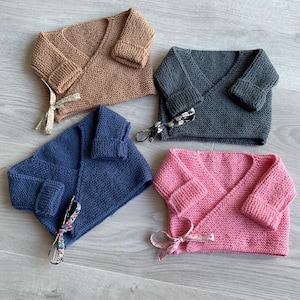 Puede incluir: Cuatro cárdigans envolventes de punto para bebé en tonos marrón, gris, azul y rosa. Cada cárdigan tiene un diseño envolvente con puños doblados y una cinta decorativa. Se muestran sobre una superficie de madera clara.