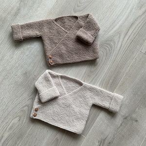 Op de afbeelding: Twee gebreide babytruien in beige en wit met houten knopen. De truien liggen op een licht houten oppervlak.