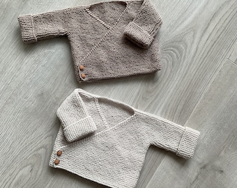 May include: Two knitted baby sweaters in beige and white with wooden buttons. The sweaters are laid out on a light wood surface.