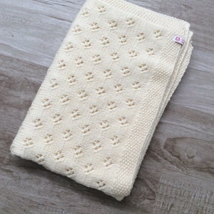 May include: A cream-colored knitted baby blanket with a delicate openwork pattern. The blanket is folded in half and has a small pink label with the text "Knitted with Love" on it.
