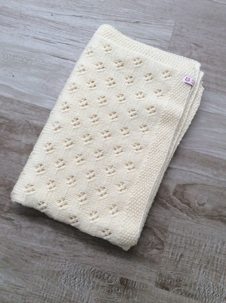 May include: A cream-colored knitted baby blanket with a delicate openwork pattern. The blanket is folded in half and has a small pink label with the text "Knitted with Love" on it.