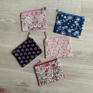 Può includere: Cinque piccole pochette quadrate in tessuto con cerniere e ciondoli in metallo. Le pochette presentano motivi floreali in vari colori, tra cui rosa, blu e viola, su sfondi blu navy, bianchi e rosa.