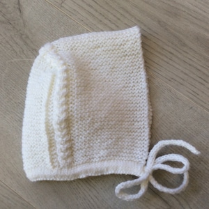 May include: A white knitted baby bonnet with a braided detail and a tie at the back.