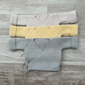 Op de afbeelding: Drie gebreide babytruien in grijs, geel en beige. De truien zijn op elkaar gevouwen en hebben een wikkel-stijl met banden.