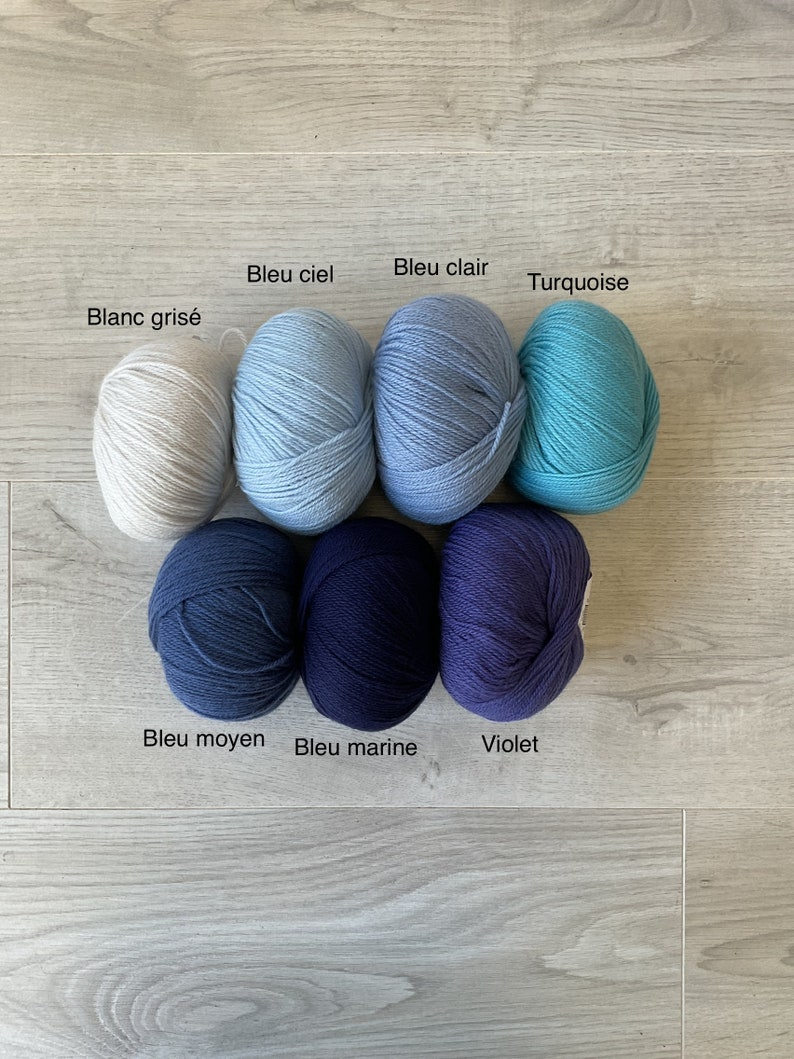 May include: Seven balls of yarn in various shades of blue, from white to dark blue, with one ball of white yarn. The yarn balls are labeled with their color names in French: Blanc gris&eacute;, Bleu ciel, Bleu clair, Turquoise, Bleu moyen, Bleu marine, and Violet.