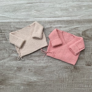 Puede incluir: Dos jerséis de punto para bebé, uno de color beige claro y el otro de color rosa pálido. Ambos jerséis tienen mangas largas y un estilo envolvente.