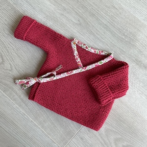 Puede incluir: Un jersey de punto rojo para bebé con ribete de estampado floral. El jersey tiene un cierre de envoltura y manga larga.