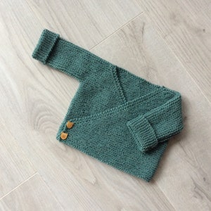 Op de afbeelding: Een groene gebreide babytrui met een wikkel sluiting en twee houten knopen.