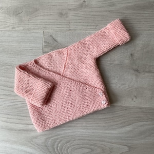 Op de afbeelding: Een roze gebreide baby-trui met knoopsluiting. De trui heeft lange mouwen en een simpel ontwerp.