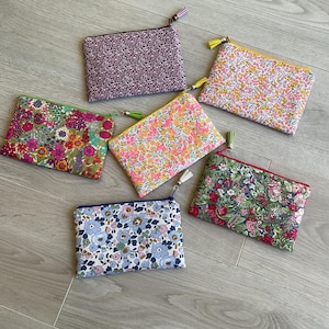 May include: Six rectangular zippered pouches featuring various floral patterns in shades of purple, pink, yellow, green, and blue. Each pouch includes a small decorative tassel attached to the zipper pull, showcasing distinct fabric designs.