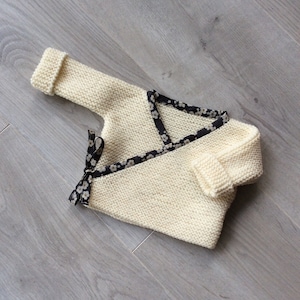 Puede incluir: Un jersey de punto crema para bebé con un ribete de cinta floral blanco y negro. El jersey tiene un estilo envolvente y se ata en la parte delantera.