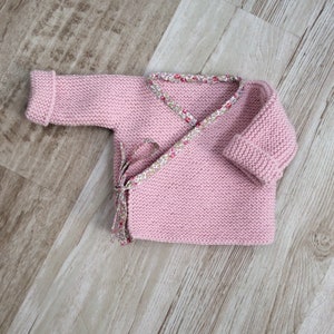 Puede incluir: Un jersey de punto rosa para bebé con un ribete floral alrededor del cuello y un cierre de lazo. El jersey está colocado plano sobre una superficie de madera.