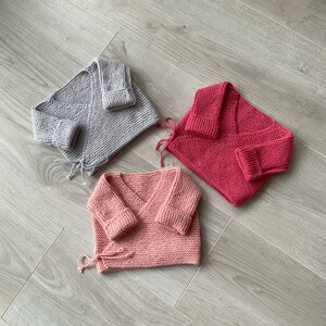 Op de afbeelding: Drie gebreide babytruien in verschillende kleuren: grijs, roze en rood. De truien liggen op een houten vloer.