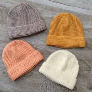 Peut inclure: Quatre bonnets en tricot de différentes couleurs : gris, jaune moutarde, pêche et blanc. Les bonnets sont disposés sur une surface en bois.