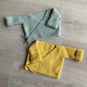 Op de afbeelding: Twee gebreide baby-vestjes, één in lichtgroen en één in geel. Beide vestjes hebben een wikkel-stijl en worden in de taille vastgebonden.