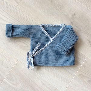 Puede incluir: Un jersey de punto azul para bebé con ribete de tela con estampado blanco y azul. El jersey tiene un estilo envolvente con cierre de nudo.