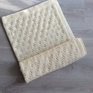 May include: A cream-colored knitted baby blanket with a delicate diamond pattern. The blanket is folded in half and is lying on a light gray wooden surface.