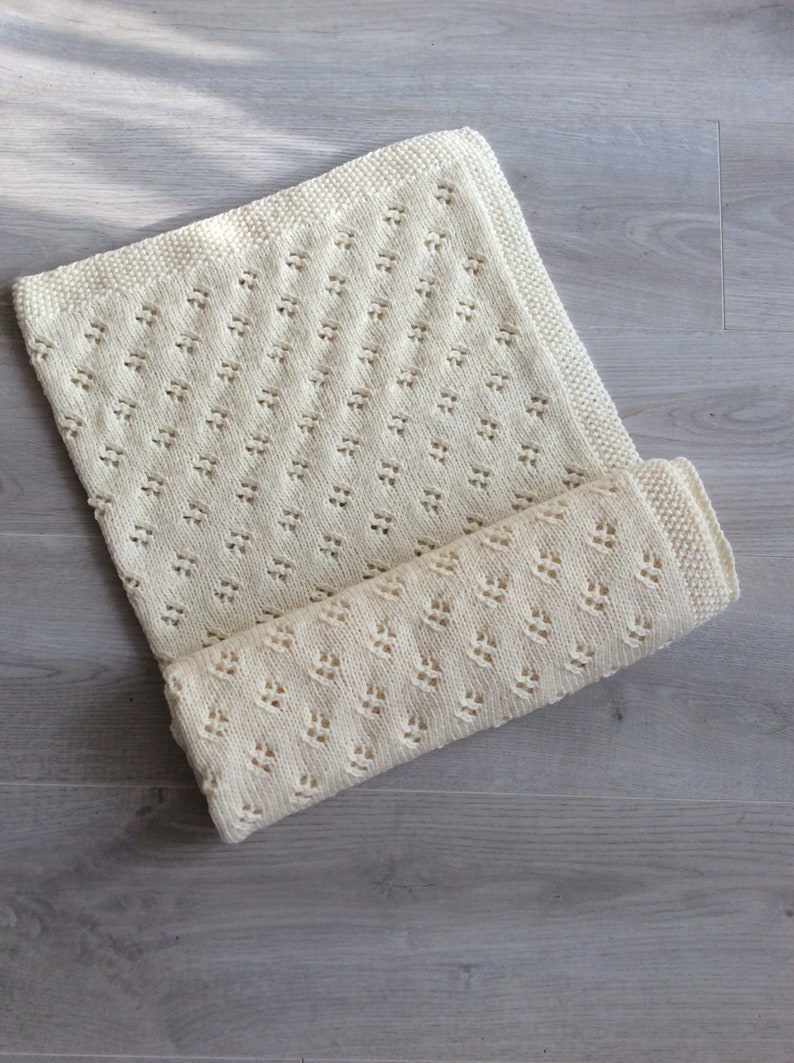 May include: A cream-colored knitted baby blanket with a delicate diamond pattern. The blanket is folded in half and is lying on a light gray wooden surface.