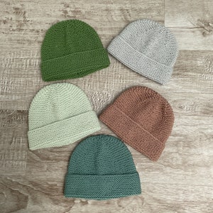 Peut inclure: Cinq bonnets en tricot de différentes couleurs : vert, vert clair, marron, gris et sarcelle. Les bonnets sont tous du même style, avec une forme simple et arrondie.