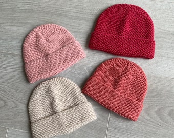 May include: Four knitted hats in different shades of pink and beige. The hats are all the same style, with a simple, rounded crown and a folded brim.
