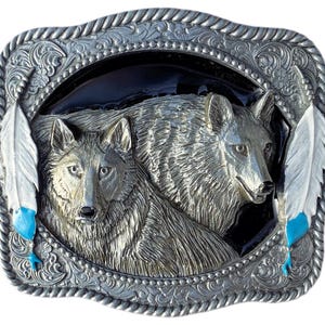 May include: Silver-toned metal belt buckle featuring two detailed wolf figures within an oval frame. The buckle has a decorative border with feather accents in white and turquoise. The overall design is intricate and detailed.