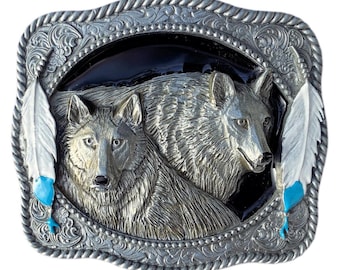May include: Silver-toned metal belt buckle featuring two detailed wolf figures within an oval frame. The buckle has a decorative border with feather accents in white and turquoise. The overall design is intricate and detailed.